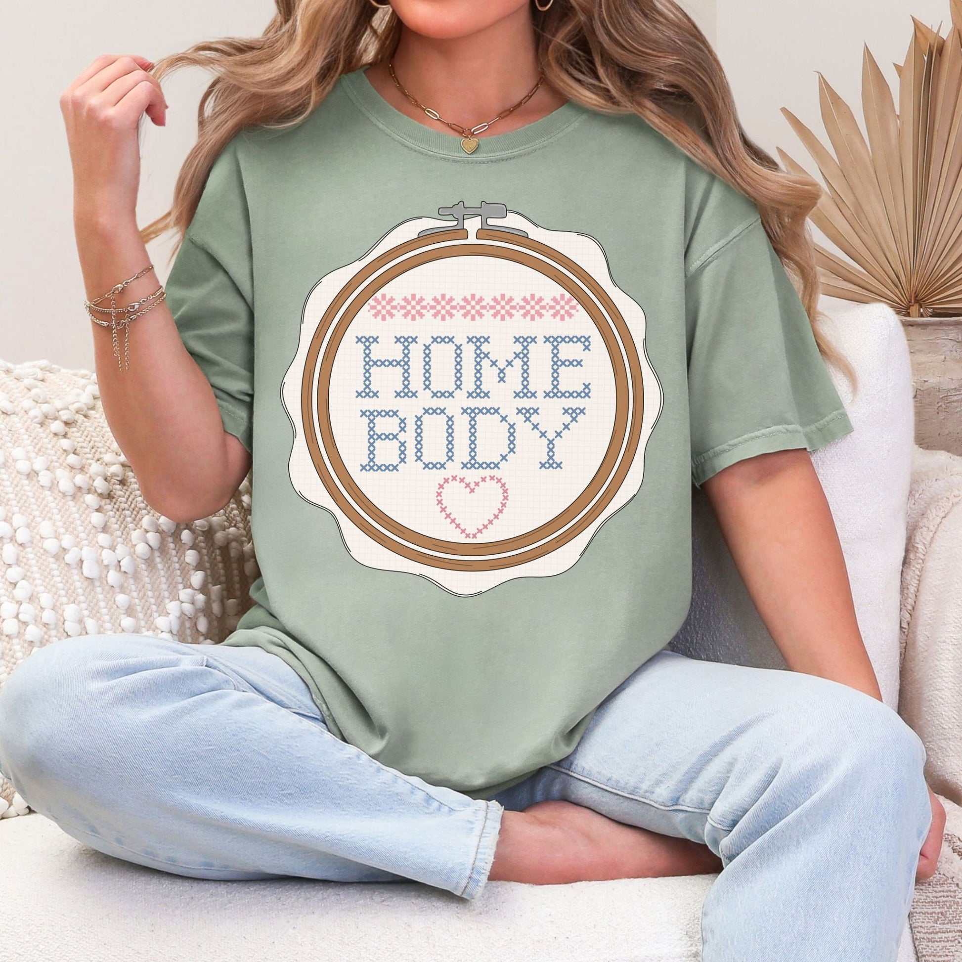 “Embroidery Hoop Home Body" graphic on muted teal-green (bay) t-shirt.”