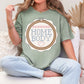 “Embroidery Hoop Home Body" graphic on muted teal-green (bay) t-shirt.”