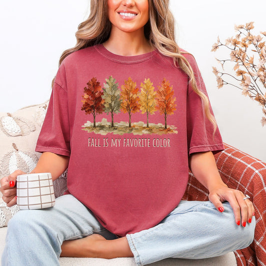 Model wearing a Comfort Colors® t-shirt in muted dark red (brick) featuring a row of five autumn trees in red, green, orange, and golden yellow with the words “Fall Is My Favorite Color.”