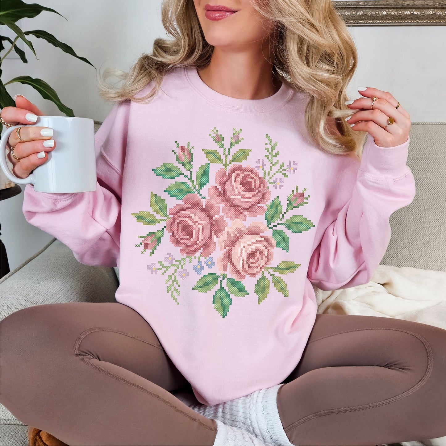 Model wearing a soft pale pink (light pink) Gildan 18000 Sweatshirt featuring a watercolor illustration designed to resemble faux cross stitch, showing a bouquet of soft pink roses with green leaves and small floral accents. The design is printed artwork and not real stitching or embroidery.