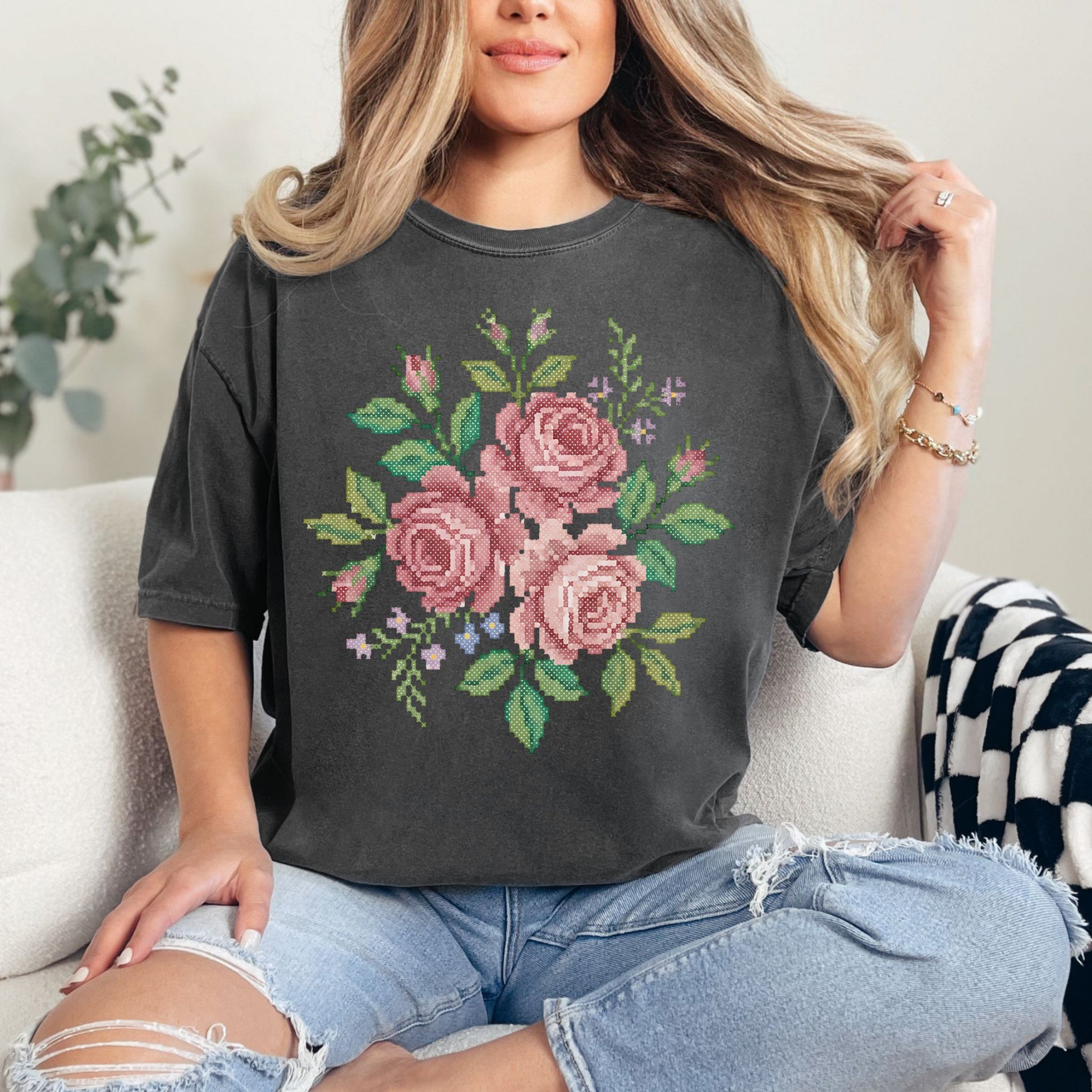 Model wearing a dark charcoal gray (pepper) Comfort Colors 1717 T-Shirt featuring a watercolor illustration designed to resemble faux cross stitch, showing a bouquet of soft pink roses with green leaves and small floral accents. The design is printed artwork and not real stitching or embroidery.