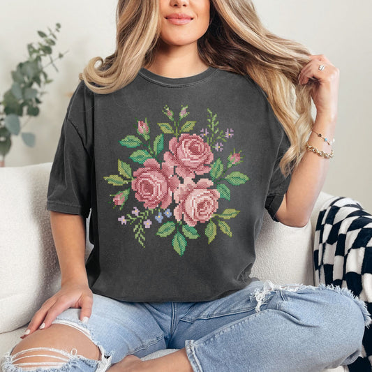 Model wearing a dark charcoal gray (pepper) Comfort Colors 1717 T-Shirt featuring a watercolor illustration designed to resemble faux cross stitch, showing a bouquet of soft pink roses with green leaves and small floral accents. The design is printed artwork and not real stitching or embroidery.