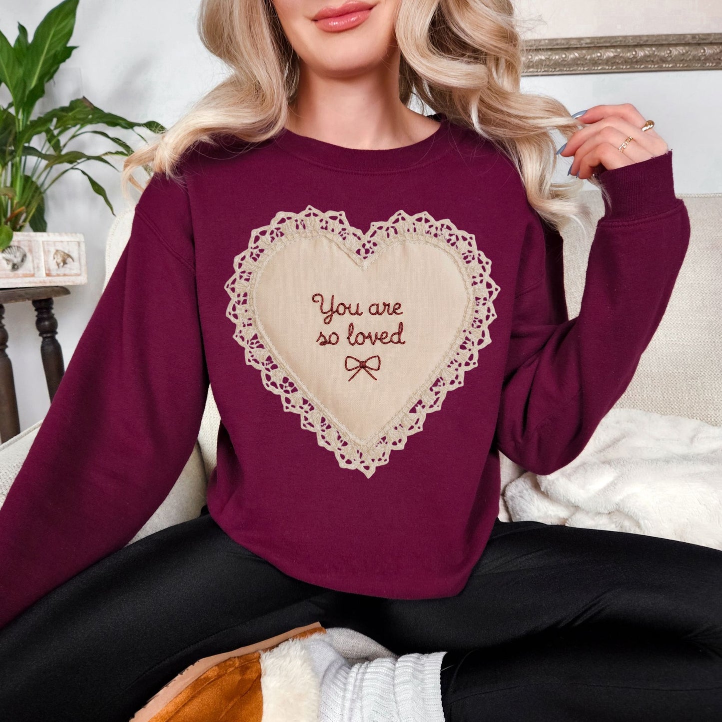 Model wearing a Gildan 18000 deep burgundy red (maroon)t-shirt featuring a lace-style heart design with the words “You are so loved” and a small bow in the center. The design resembles embroidery stitching but is a printed illustration. Shown on a deep red (chili) Comfort Colors shirt.
