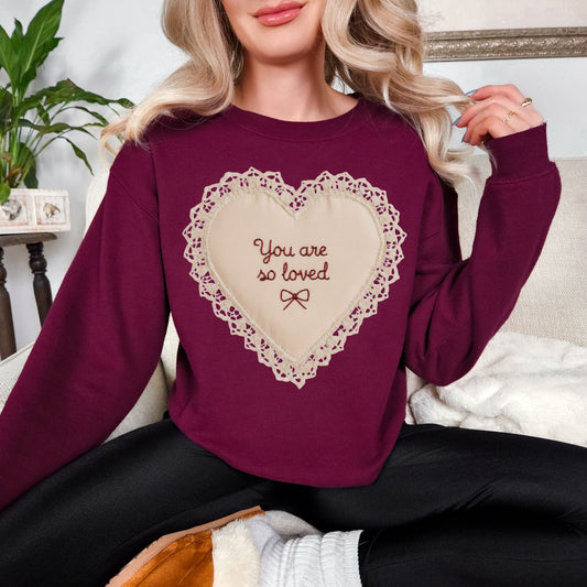 Model wearing a Gildan 18000 deep burgundy red (maroon)t-shirt featuring a lace-style heart design with the words “You are so loved” and a small bow in the center. The design resembles embroidery stitching but is a printed illustration. Shown on a deep red (chili) Comfort Colors shirt.