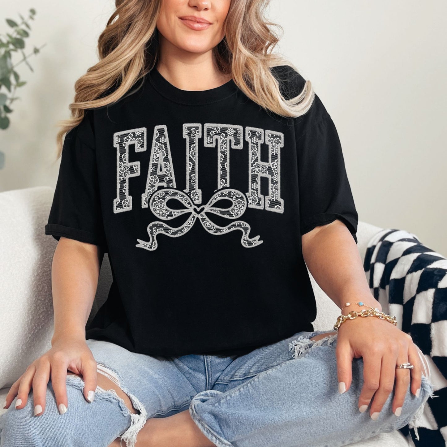 Model wearing a Comfort Colors 1717 Black T-shirt featuring the word “FAITH” in large faux embroidery-style letters filled with delicate lace patterns, with a decorative bow beneath the text. The design is printed artwork made to resemble embroidered lace stitching but is not real embroidery or textured thread.
