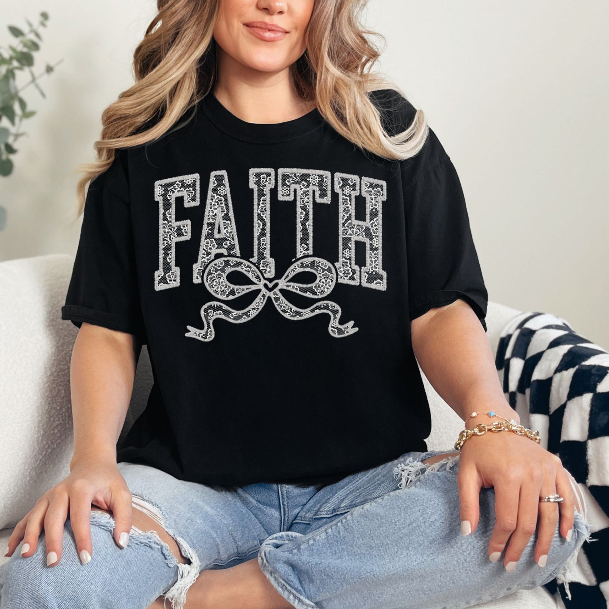 Model wearing a Comfort Colors 1717 Black T-shirt featuring the word “FAITH” in large faux embroidery-style letters filled with delicate lace patterns, with a decorative bow beneath the text. The design is printed artwork made to resemble embroidered lace stitching but is not real embroidery or textured thread.