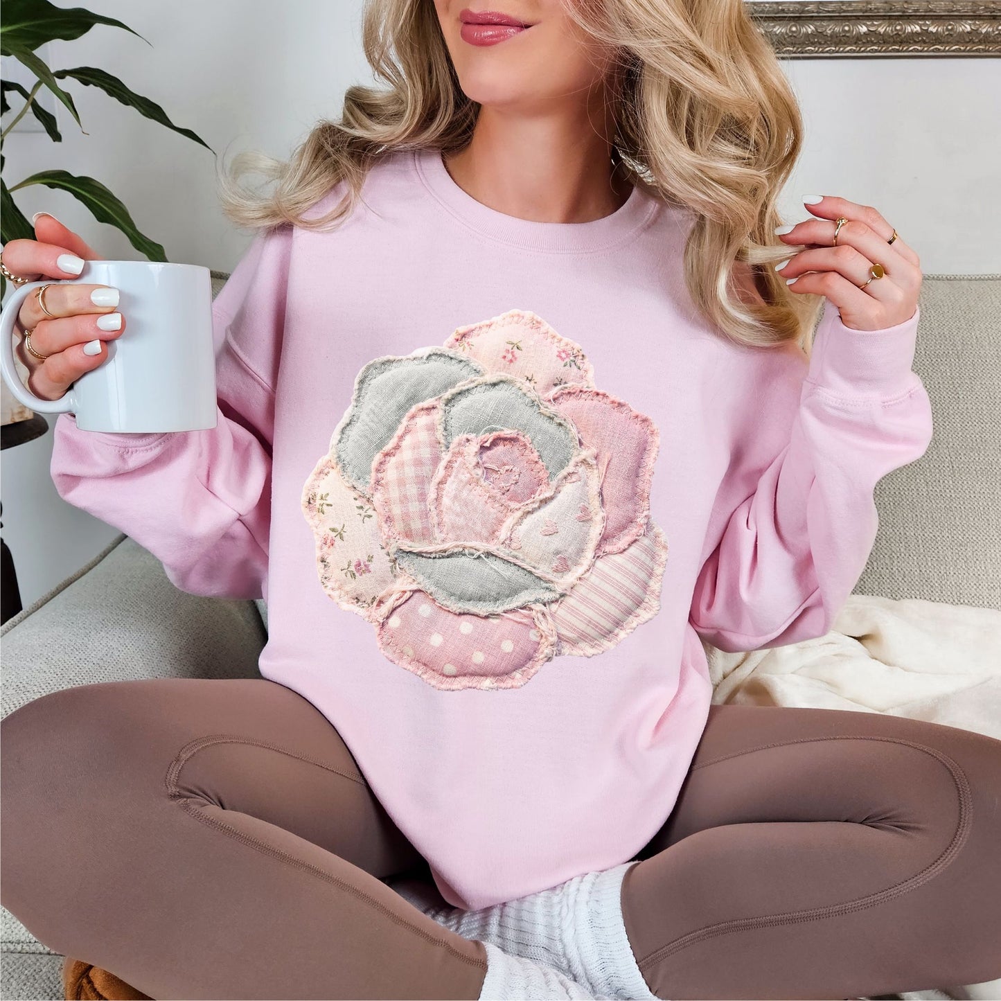 Model wearing Gildan 18000 Sweatshirt in soft pale pink (light pink) featuring a faux patchwork-style rose made of layered fabric-inspired patterns including florals, gingham, stripes, and polka dots in pastel pink, cream, and soft gray tones. The design is printed to resemble fabric patchwork and is not real stitched or textured material.