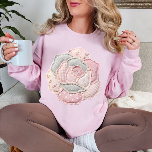 Model wearing Gildan 18000 Sweatshirt in soft pale pink (light pink) featuring a faux patchwork-style rose made of layered fabric-inspired patterns including florals, gingham, stripes, and polka dots in pastel pink, cream, and soft gray tones. The design is printed to resemble fabric patchwork and is not real stitched or textured material.