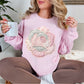 Model wearing Gildan 18000 Sweatshirt in soft pale pink (light pink) featuring a faux patchwork-style rose made of layered fabric-inspired patterns including florals, gingham, stripes, and polka dots in pastel pink, cream, and soft gray tones. The design is printed to resemble fabric patchwork and is not real stitched or textured material.