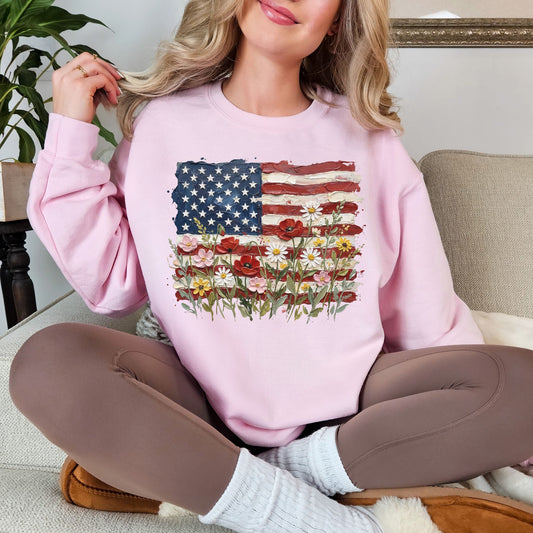 Model is wearing a Gildan 18000 sweatshirt in soft pale pink (light pink) featuring a faux painted American flag design with textured brushstroke details and colorful wildflowers blooming across the bottom.