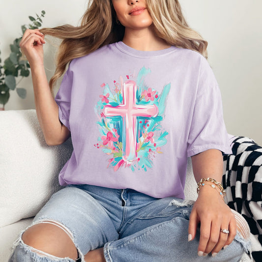 Model wearing a Comfort Colors 1717 T-Shirt in a soft purple (orchid) featuring a brushstroke graphic of a pastel pink cross with a light blue background. Darling pink flowers peak from behind the cross.