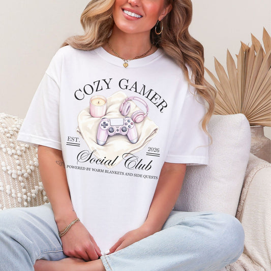 Model wearing a Comfort Colors 1717 t-shirt in a bright white (white) color featuring a cozy gamer graphic with a controller, candle and a headset. It writing above the graphic says, "Cozy Gamer". Below, it says "Social Club powered by warm blankets and side quests"
