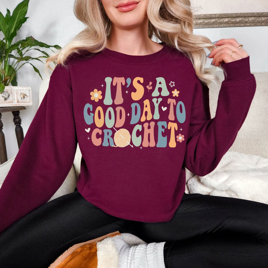 Model wearing a Gildan 18000 deep burgundy red (maroon) color featuring the words "It's a good day to crochet" in a bubble font. The letters alternate in the colors pastel green, orange- red, muted blue and an orange- yellow. Tiny flowers, hearts, and leaves surround the graphic. The "O" in crochet is an orange- red yarn ball with a crochet hook.