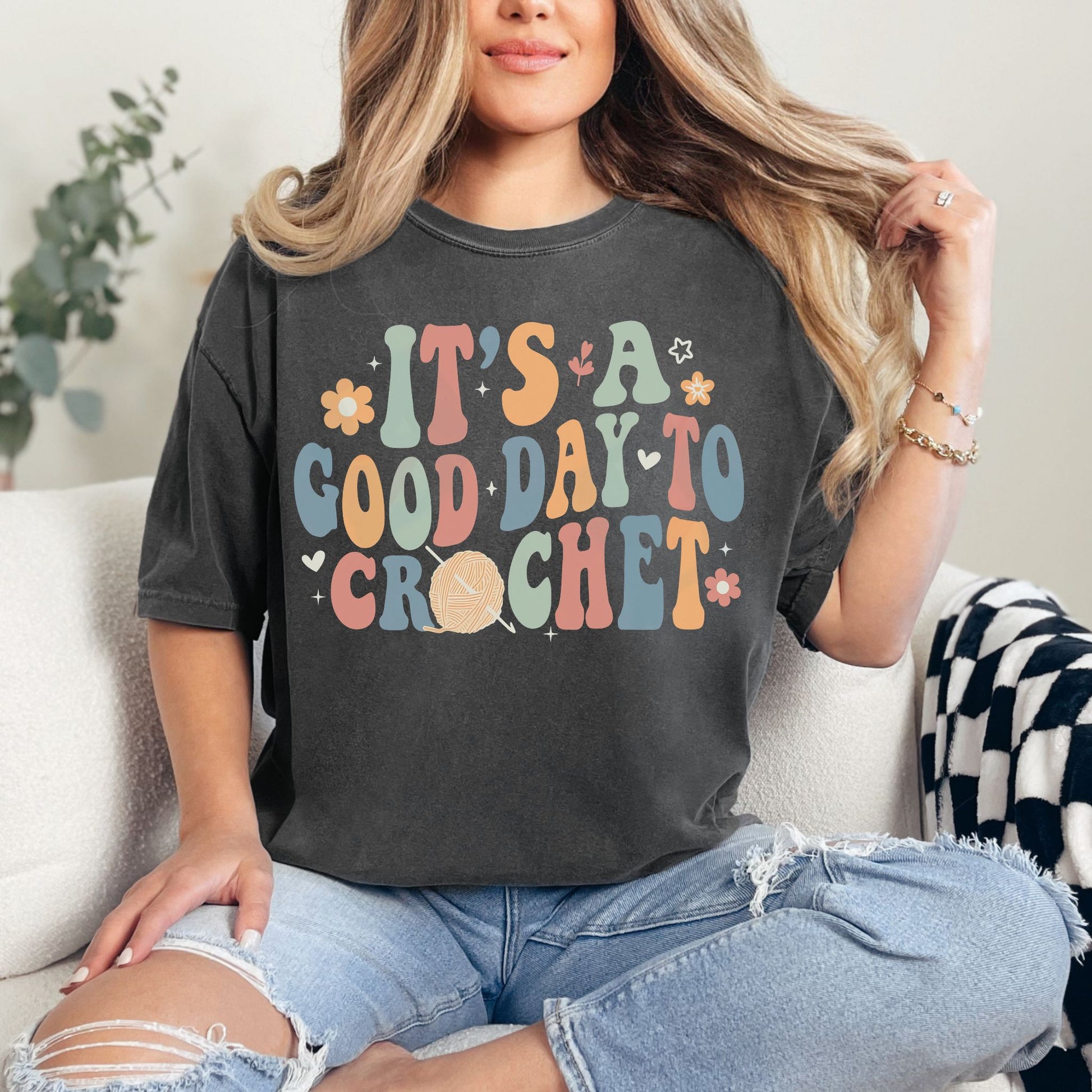 Model wearing a Comfort Colors 1717 t-shirt in a dark charcoal gray (pepper)  color featuring the words "It's a good day to crochet" in a bubble font. The letters alternate in the colors pastel green, orange- red, muted blue and an orange- yellow.  Tiny flowers, hearts, and leaves surround the graphic. The "O" in crochet is an orange- red yarn ball with a crochet hook.