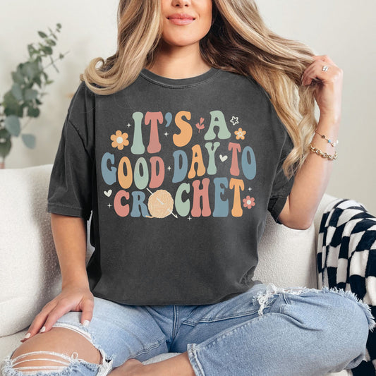 Model wearing a Comfort Colors 1717 t-shirt in a dark charcoal gray (pepper) color featuring the words "It's a good day to crochet" in a bubble font. The letters alternate in the colors pastel green, orange- red, muted blue and an orange- yellow. Tiny flowers, hearts, and leaves surround the graphic. The "O" in crochet is an orange- red yarn ball with a crochet hook.