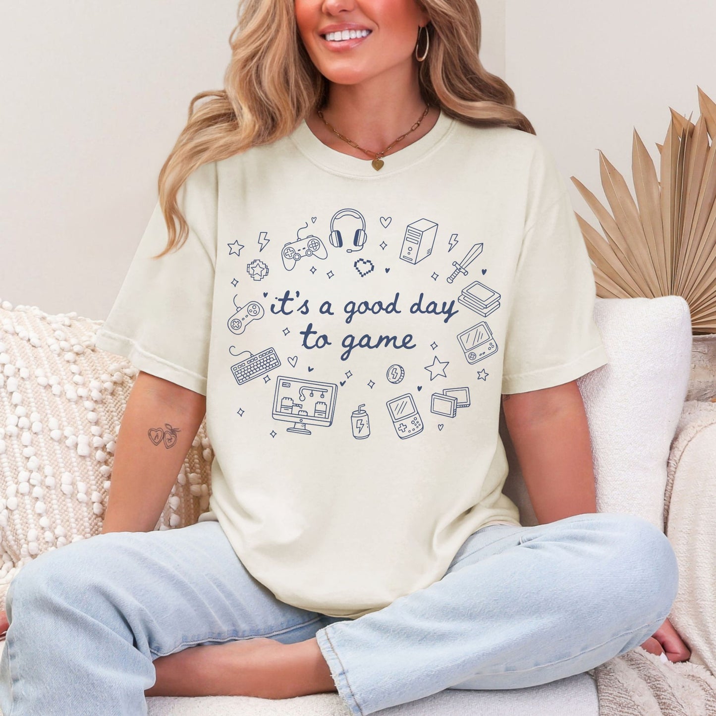 Model wearing a Comfort Colors 1717 t-shirt in a soft off-white (ivory) color featuring doodle illustrations of gaming products. For example, controllers, headset, and game consoles. Other elements are hearts and stars. In the middle of the gaming doodles is the phrase "It's a good day to game" in a hand-written cursive type of font. 
