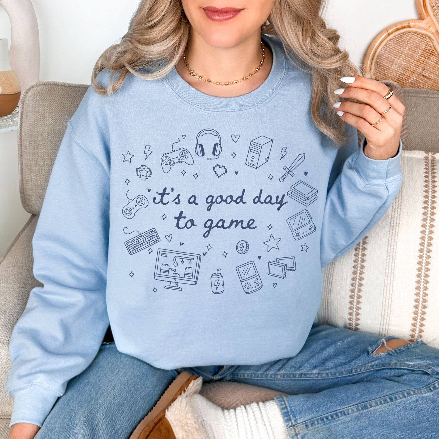 Model wearing a Gildan 18000 Sweatshirt in pale soft blue (light blue) color featuring doodle illustrations of gaming products. For example, controllers, headset, and game consoles. Other elements are hearts and stars. In the middle of the gaming doodles is the phrase "It's a good day to game" in a hand-written cursive type of font. 