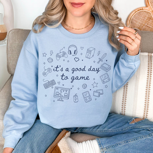 Model wearing a Gildan 18000 Sweatshirt in pale soft blue (light blue) color featuring doodle illustrations of gaming products. For example, controllers, headset, and game consoles. Other elements are hearts and stars. In the middle of the gaming doodles is the phrase "It's a good day to game" in a hand-written cursive type of font. 