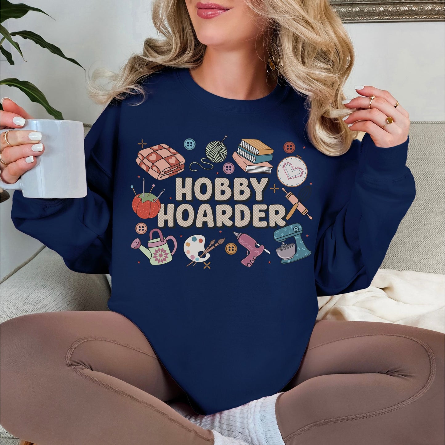 Model wearing a Gildan 18000 sweatshirt in deep navy blue (navy) color featuring illustrations of hobbies such as blankets, watering can, books and painting supplies with the words "Hobby Hoarder" in the middle in a bubble type font.