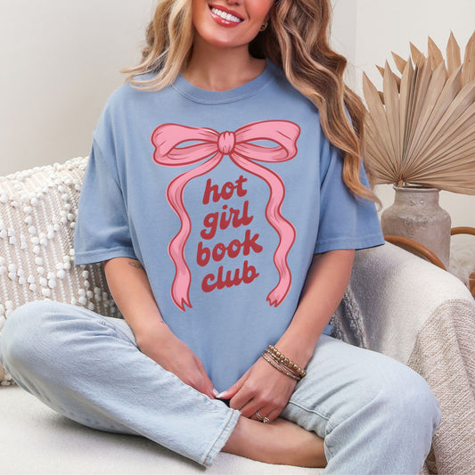 “Model wearing a Comfort Colors 1717 t-shirt in muted denim blue (washed denim) color featuring a graphic design of a large pink bow with the phrase "hot girl book club" underneath in between the ribbons.