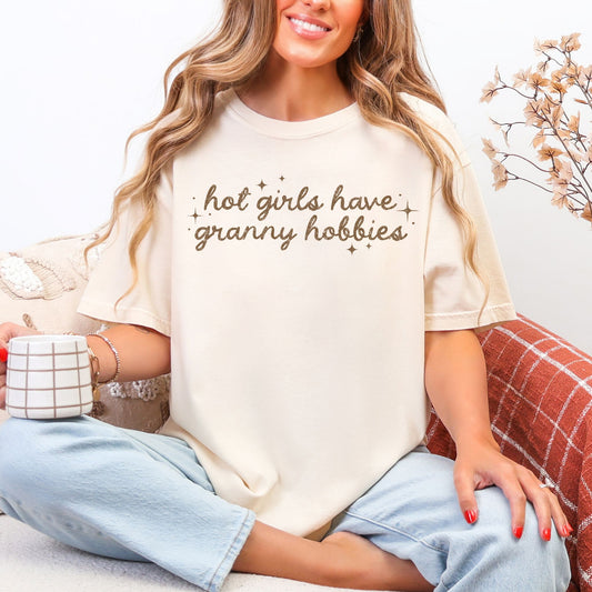Model wearing a Comfort Colors® 1717 short sleeve t-shirt in soft cream (ivory) featuring a neutral brown script typography design that reads “Hot Girls Have Granny Hobbies” surrounded by small decorative sparkle stars.