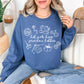 Model wearing a Gildan 18000 Sweatshirt in a rich medium-dark blue (indigo blue) color featuring cute sewing hobby line art illustrations, which include a sewing machine, an embroidery hoop and knitting needles with a blanket attached. In the center of the illustrations is the phrase "hot girls have grandma hobbies" in a hand-written script font.