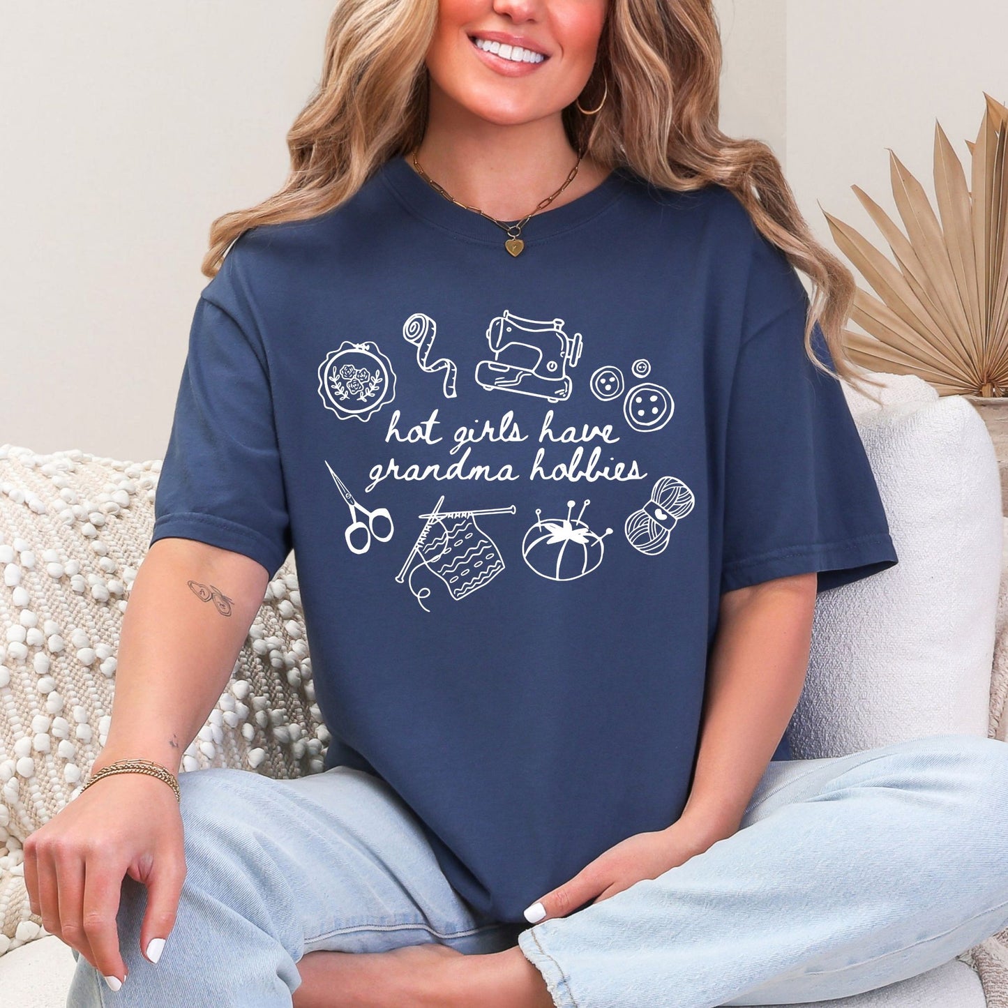 Model wearing a Comfort Colors 1717 t-shirt in a deep navy blue (navy) color featuring cute sewing hobby line art illustrations, which include a sewing machine, an embroidery hoop and knitting needles with a blanket attached. In the center of the illustrations is the phrase "hot girls have grandma hobbies" in a hand-written script font.