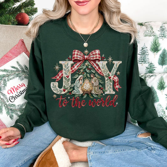 Model wearing a Gildan 18000 Long sleeve sweatshirt in dark green (forest green) featuring a Christmas design with the words “JOY to the world,” a nativity scene beneath an evergreen tree, a red and cream plaid bow, greenery, red berries, and gold stars.