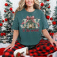 Model wearing a Comfort Colors® 1717 short-sleeve t-shirt in dark blue-green (Blue Spruce) featuring a Christmas design with the words “JOY to the world,” a nativity scene beneath an evergreen tree, a red and cream plaid bow, greenery, red berries, and gold stars.