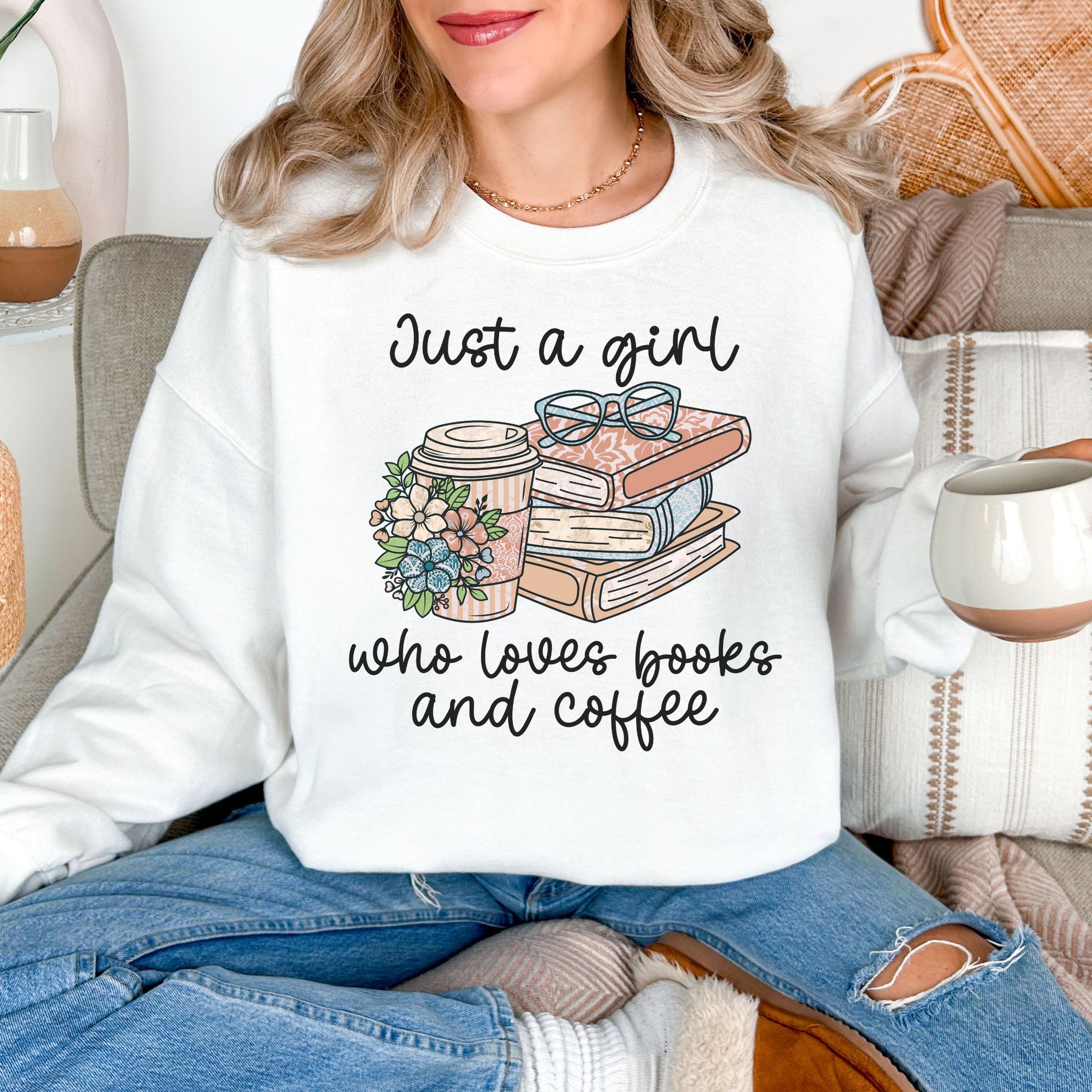 Model wearing a Gildan 18000 Sweatshirt in a clean bright white (white) featuring a neutral-colored graphic design of a stack of books, a to go coffee, a pair of eyeglasses and florals. The cursive script reads "Just a girl who loves books and coffee".