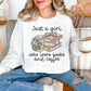 Model wearing a Gildan 18000 Sweatshirt in a clean bright white (white) featuring a neutral-colored graphic design of a stack of books, a to go coffee, a pair of eyeglasses and florals. The cursive script reads "Just a girl who loves books and coffee".