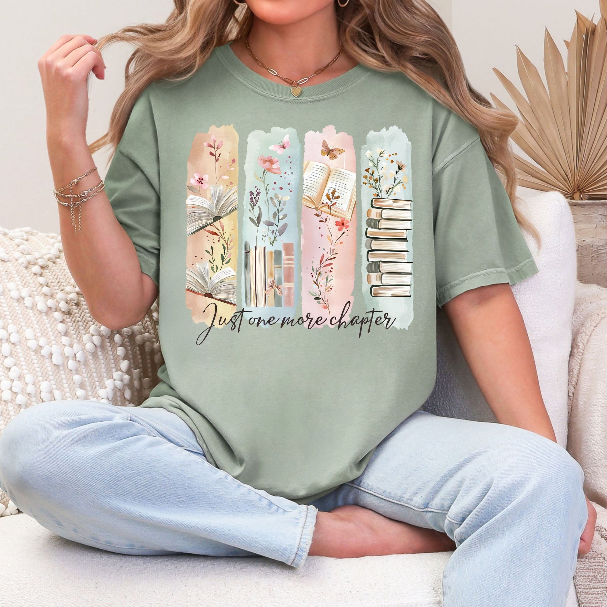 Model is wearing a Soft green (bay) Comfort Colors 1717 T-shirt featuring a watercolor-style illustration of vertical panels with open books, stacked books, florals, and delicate nature accents in muted pastel tones. The script text below reads “Just one more chapter.”