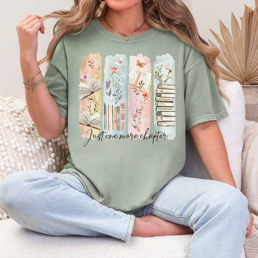 Model is wearing a Soft green (bay) Comfort Colors 1717 T-shirt featuring a watercolor-style illustration of vertical panels with open books, stacked books, florals, and delicate nature accents in muted pastel tones. The script text below reads “Just one more chapter.”