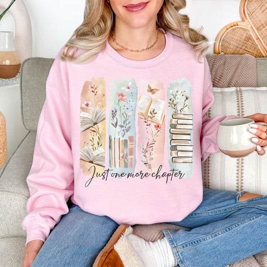 Model is wearing a soft pale pink (light pink) Gildan 18000 Sweatshirt featuring a watercolor-style illustration of vertical panels with open books, stacked books, florals, and delicate nature accents in muted pastel tones. The script text below reads “Just one more chapter.”
