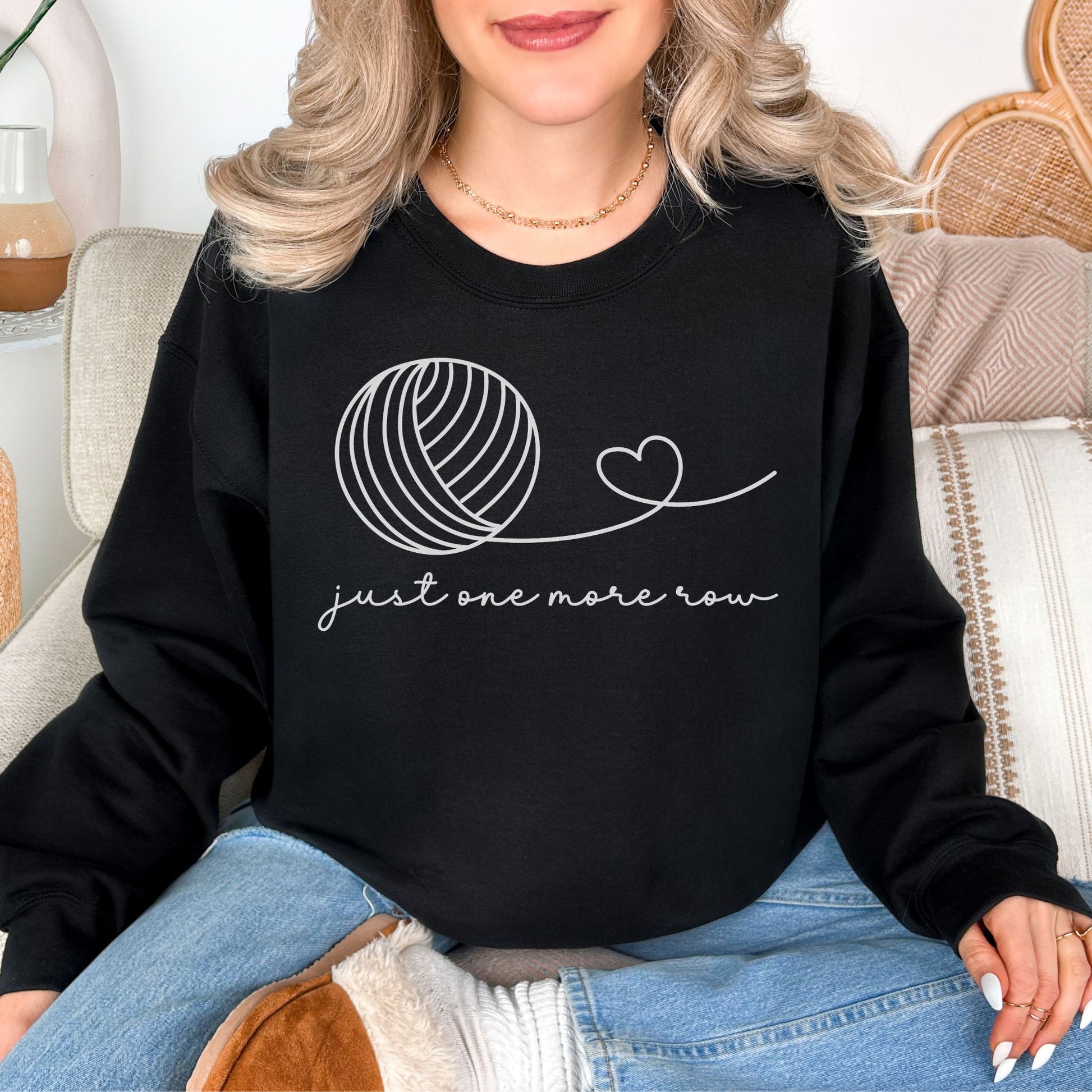 Model wearing a Gildan 18000 Sweatshirt in a deep black (black) color featuring a line art yarn ball with a string from the ball that turns into a heart. Below the image is the phrase "just one more row" in a cursive font.