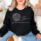 Model wearing a Gildan 18000 Sweatshirt in a deep black (black) color featuring a line art yarn ball with a string from the ball that turns into a heart. Below the image is the phrase "just one more row" in a cursive font.