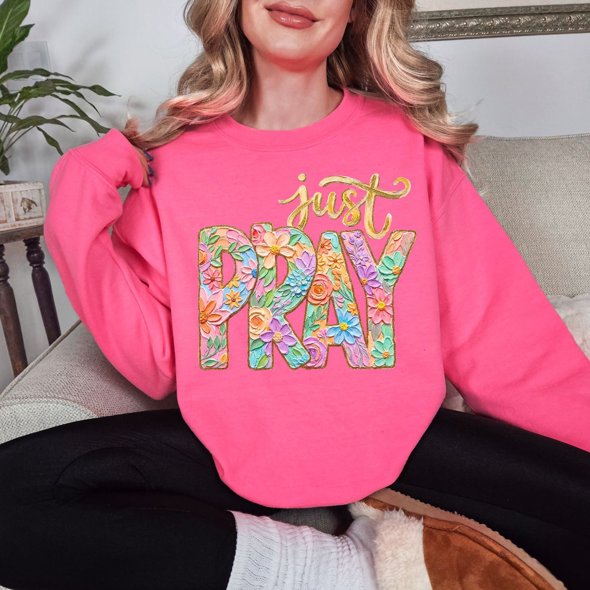 Model wearing a Gildan 18000 fluorescent bright pink (safety pink) sweatshirt featuring a floral “Just Pray” Christian graphic with pastel flowers inside bold lettering and gold script text above, designed in a faux painted style.