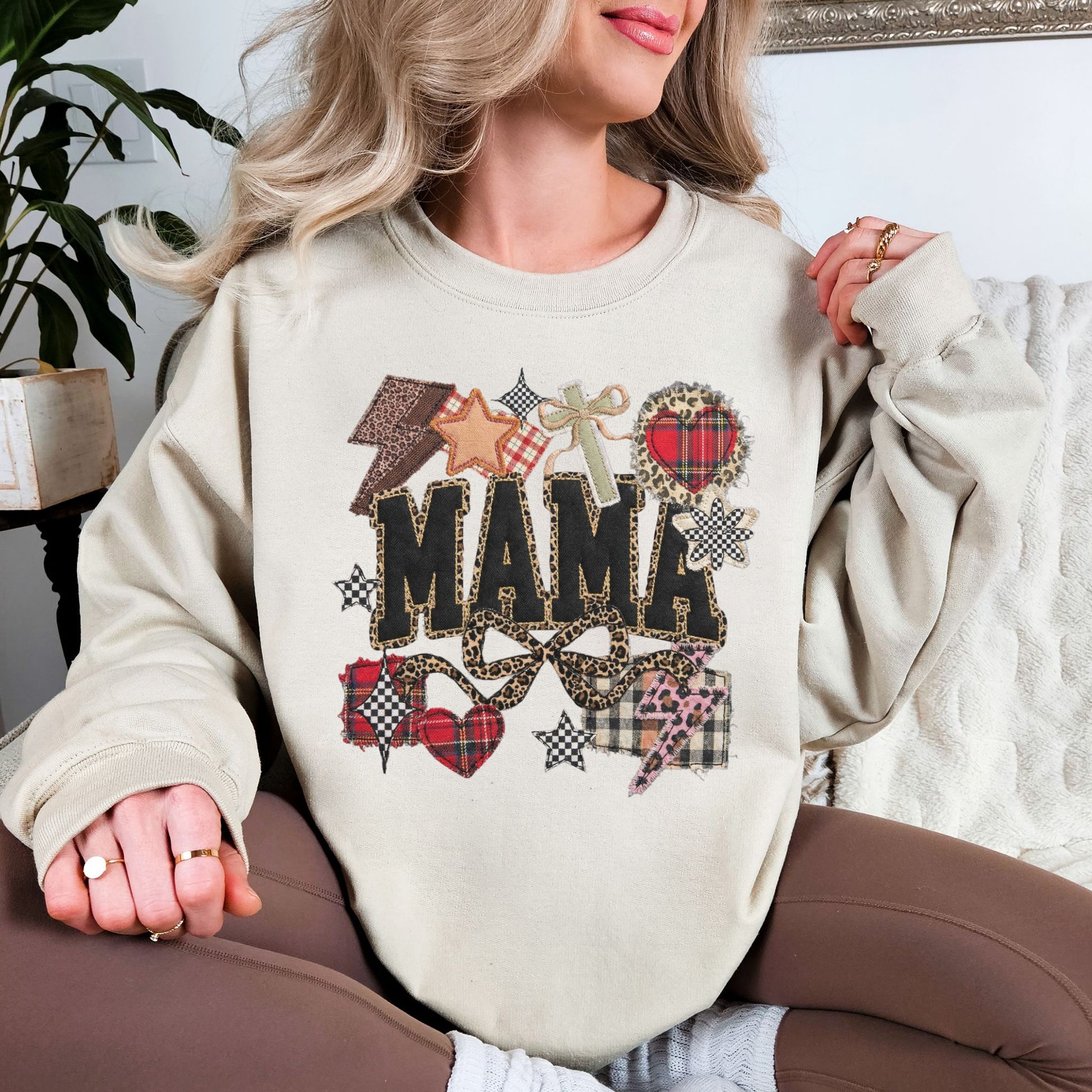 Model is wearing a Gildan 18000 light beige (sand) sweatshirt featuring the word “mama” in a faux patchwork and embroidery style with leopard print, plaid hearts, stars, and bow accents.