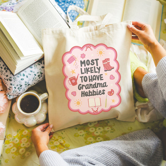 Natural canvas tote bag with "Most Likely to Have Grandma Hobbies" text held by a person on a floral surface. The design has an adorable doodle border with fun hobby images inside including a journal, watering can, and buttons with the phrase in the middle. The doodle border is in pink and red tones with dark gray to black lettering for the phrase.