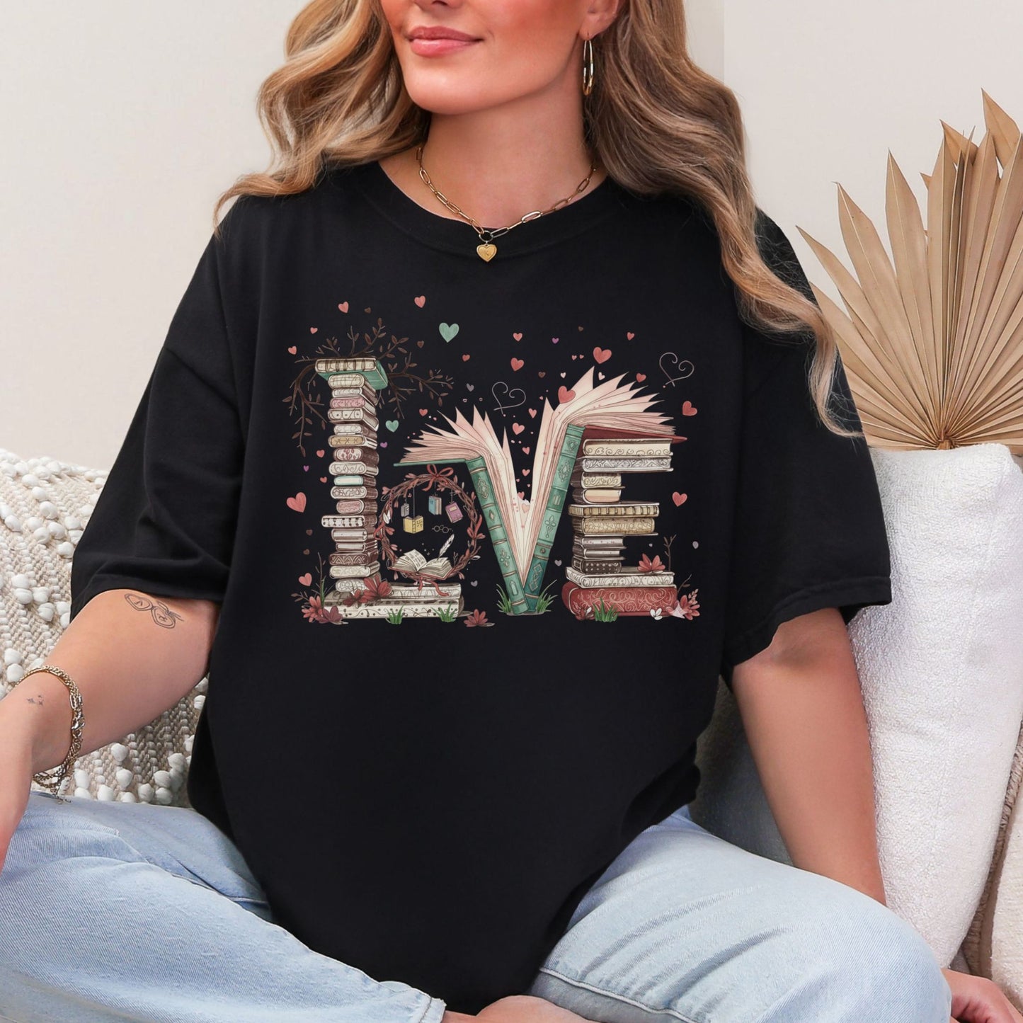 Model wearing a Comfort Colors 1717 t-shirt in black featuring a whimsical stacked book graphic that spell the word ‘Love’. Along with tiny heart and branch accents.