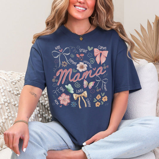 Model is wearing a Comfort Colors 1717 t-shirt in deep blue (navy) featuring the word “mama” surrounded by bows, flowers, and doodle-style accents with words like strong, brave, caring, and loved in a soft feminine design.