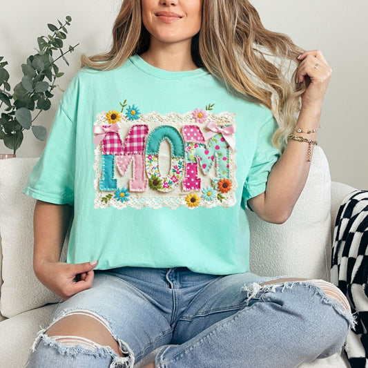 Model is wearing a Comfort Colors 1717 t-shirt in soft aqua blue (island reef) featuring the word “mom” in a floral patchwork design with bows, lace trim, and faux embroidery stitching details.