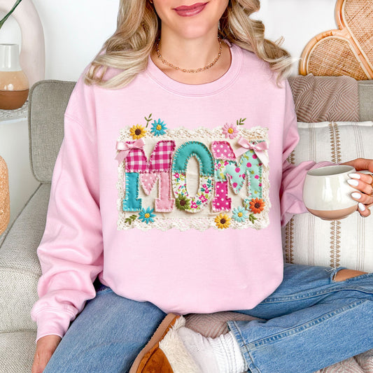 Model is wearing a Gildan 18000 sweatshirt in soft pale pink (light pink) featuring the word “mom” in a floral patchwork design with bows, lace trim, and faux embroidery stitching details.