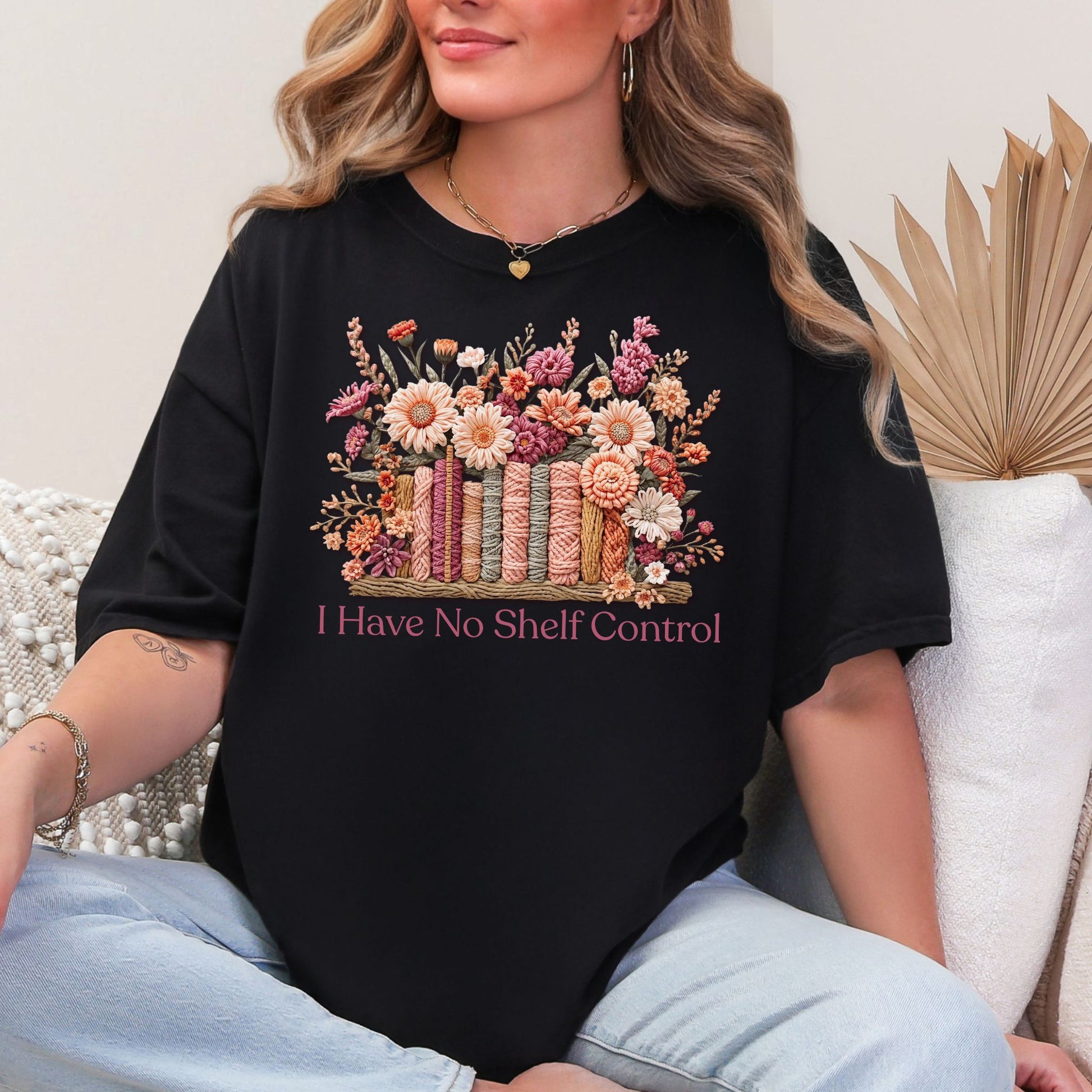 Model is wearing a Comfort Colors 1717 T-shirt in black featuring a faux yarn-style illustration of colorful books wrapped in textured thread, topped with floral arrangements in soft pink, peach, and muted green tones. The text below reads “I Have No Shelf Control."
