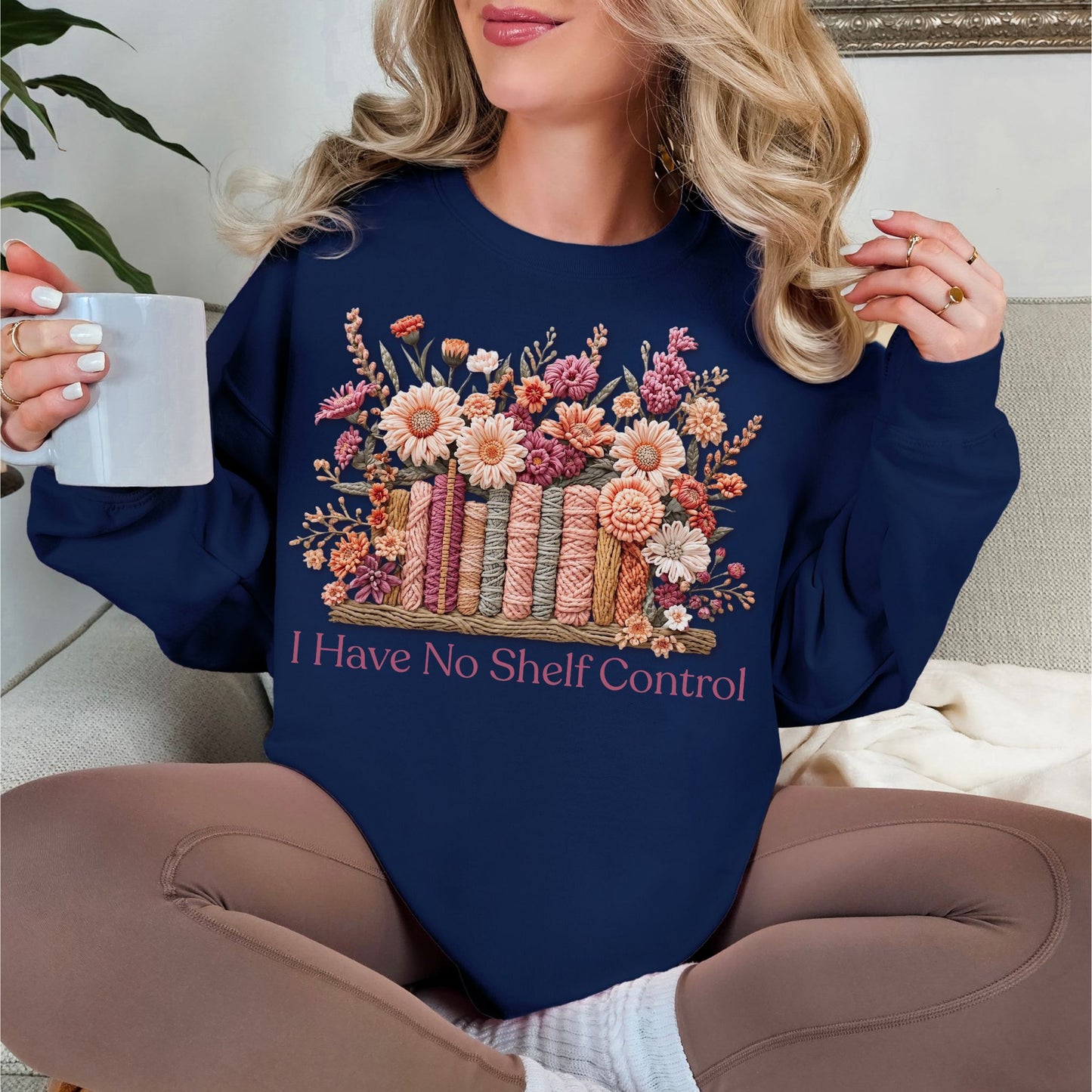 Model is wearing a Gildan 18000 Sweatshirt in deep navy blue (navy) featuring a faux yarn-style illustration of colorful books wrapped in textured thread, topped with floral arrangements in soft pink, peach, and muted green tones. The text below reads “I Have No Shelf Control."
