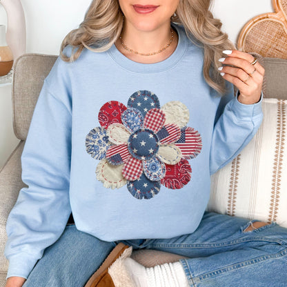 Model wearing a Gildan 18000 sweatshirt in blue (light blue) featuring a patriotic patchwork flower design with faux embroidery texture, including red, white, and blue bandana prints, floral patterns, and striped fabric details.
