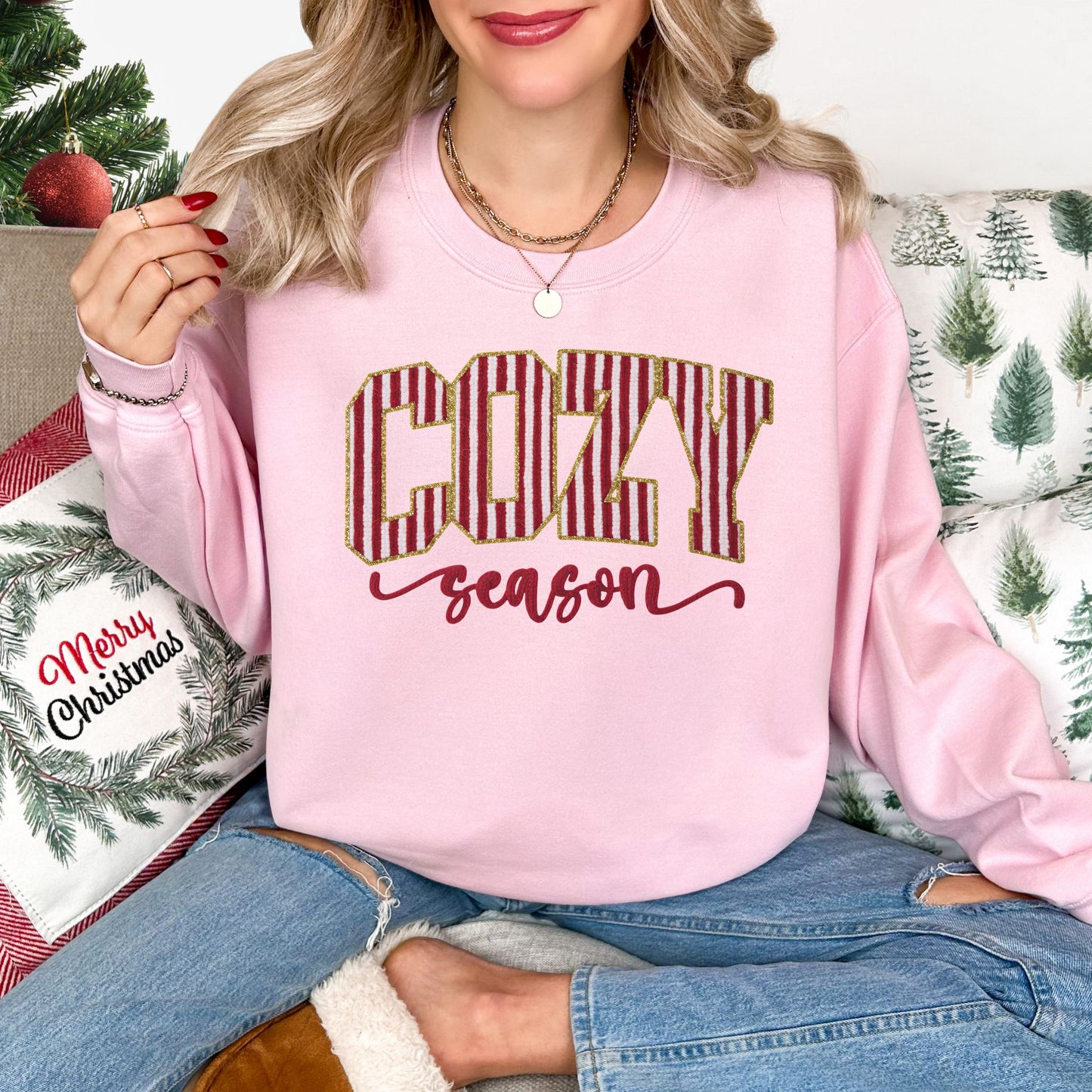 Model wearing a Gildan 18000 crewneck sweatshirt in pale pink (Light Pink) featuring the words “COZY season,” with large red-and-white vertical peppermint-striped block letters outlined in gold-colored glitter-effect detailing and the word “season” in red cursive lettering beneath.