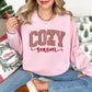 Model wearing a Gildan 18000 crewneck sweatshirt in pale pink (Light Pink) featuring the words “COZY season,” with large red-and-white vertical peppermint-striped block letters outlined in gold-colored glitter-effect detailing and the word “season” in red cursive lettering beneath.