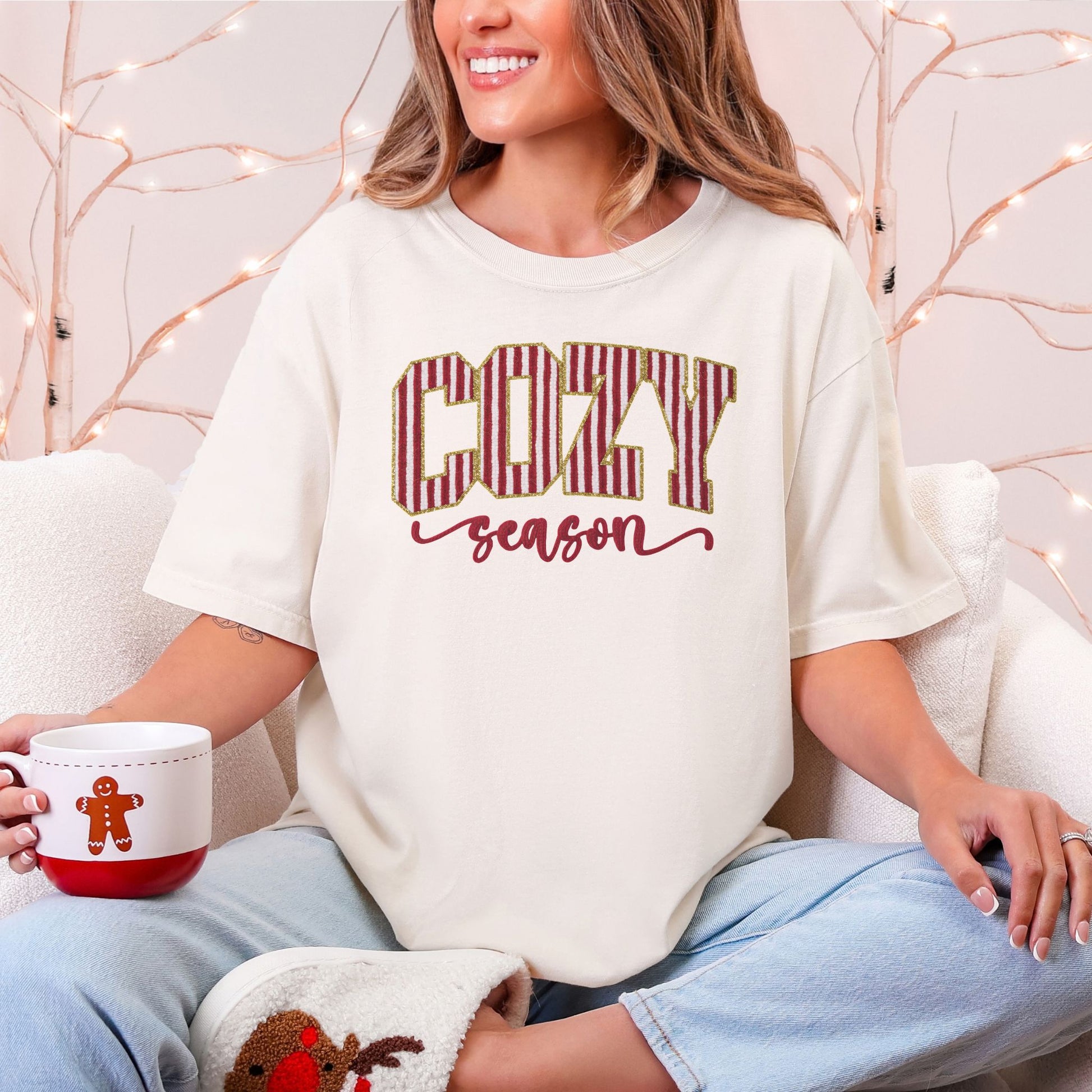 Model wearing a Comfort Colors 1717 T-shirt in soft off-white (ivory) featuring the words “COZY season,” with large red-and-white vertical peppermint-striped block letters outlined in gold-colored glitter-effect detailing and the word “season” in red cursive lettering beneath.