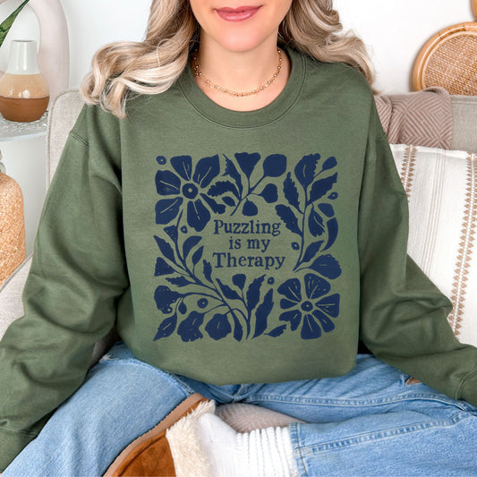 Model is wearing a Gildan 18000 muted olive green (military green) sweatshirt. The shirt has a floral dark blue design that surrounds the words "Puzzling is my therapy".